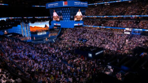 A large crowd attends a political event inside an arena. A person speaks on a stage, with their image displayed on multiple large screens. Banners and signs display "DNC 2020" and "Build Back Better.