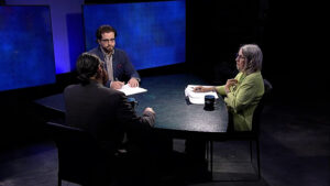 Three people are seated around a table during a televised discussion, with two large screens in the background.