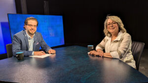 Two people are sitting at a table in a studio setting, one wearing a blue suit and the other in a white blouse. Both have coffee mugs in front of them. A large screen is visible in the background.