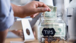 A hand places a dollar bill into a clear glass jar labeled "TIPS," which is filled with various bills and coins.