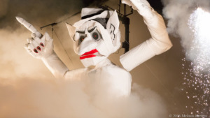 A large, white puppet with exaggerated facial features and red lips is surrounded by smoke and fireworks during a nighttime event.