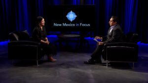 Two people are seated and conversing on a set with a dark blue backdrop, in front of a screen displaying "New Mexico in Focus.