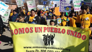 A group of people, including children, hold signs and banners that say "SOMOS UN PUEBLO UNIDO" and messages supporting parents, during a rally in Santa Fe, NM.