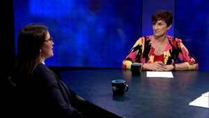 Two women are engaged in a conversation at a round table on a set with a blue background. One woman is facing the camera, and the other has her back to it. Cups and papers are on the table.