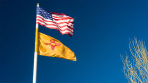 An American flag flies above a New Mexico state flag on a flagpole against a clear blue sky.