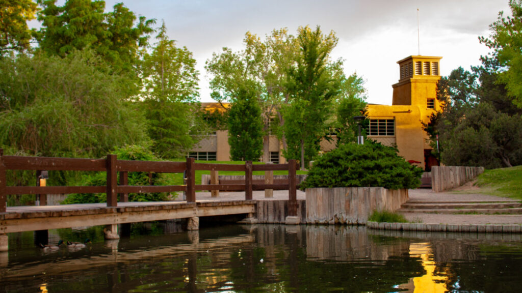 A wooden bridge crosses a pond with calm water, leading to a building surrounded by green trees and bushes.