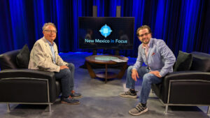 Two men sitting on a set with New Mexico in Focus logo displayed on a screen between them, facing each other on black chairs, with a blue curtain backdrop.