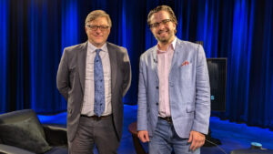 Two men stand in a studio with blue curtains. One man wears a grey suit and blue tie, while the other wears a light blue blazer and pink shirt. They are smiling at the camera.