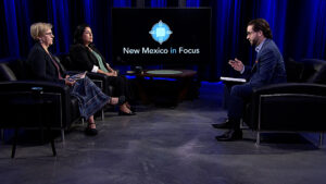 Three individuals engaged in a discussion on a television set with a large screen displaying "New Mexico in Focus" in the background. They are seated in a studio setting with blue curtains.