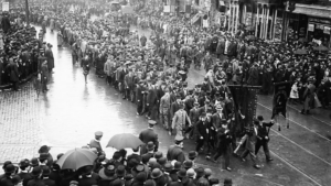 A large crowd is marching down a wet street. People are holding banners, with onlookers and bystanders on either side. The scene appears to be from an earlier time period, with everyone in formal attire.