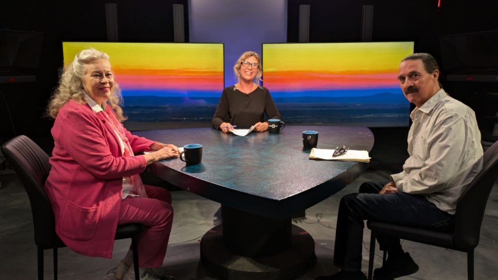 Three people sit around a table in a studio with large screens displaying a colorful sunset in the background. They have mugs and papers in front of them, and one person is holding a pen.