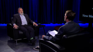 Two men are seated in armchairs in a studio with blue curtains. One man is speaking while the other, facing away, holds a notepad.