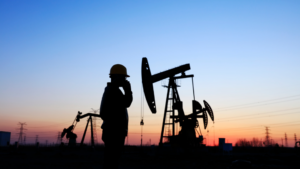 Silhouette of a worker with a hard hat talking on a phone in front of oil pump jacks during sunset.