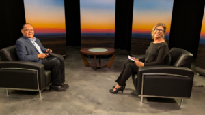 Two people are seated in armchairs on a stage with a sunset backdrop, engaged in a conversation. A small table is positioned between them.