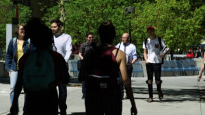 A diverse group of people walk outdoors on a sunny day, with greenery in the background. Some are carrying backpacks.