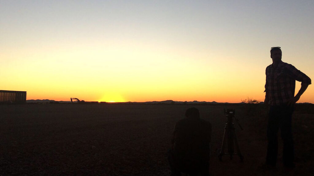 Two individuals, one standing and one crouching with a camera, watch a sunset over a barren landscape on a clear evening, their eyes reflecting the distant shimmer of water.