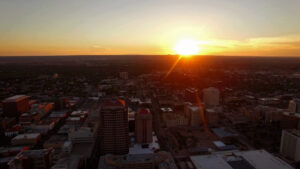 Aerial view of a cityscape at sunset, with sun setting on the horizon, casting warm light over buildings and streets.