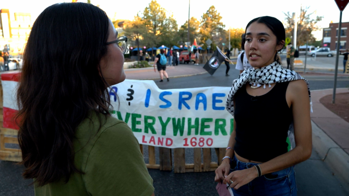 Two people converse in front of a sign at an outdoor gathering. One wears a scarf, and others are visible in the background amid trees and structures.