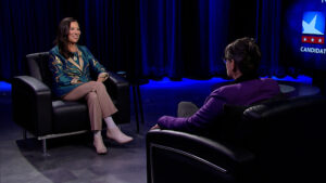 Two people sit and converse in a dimly lit studio with black chairs and curtains. A screen displays a partial logo.