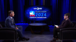 Two people seated in a studio set with "NM PBS Election 2024 Candidate Conversations" displayed on a screen between them.