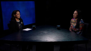 Two women sitting at a triangular table in a dimly lit studio, talking. A blue screen is in the background. Each woman has a mug in front of them.