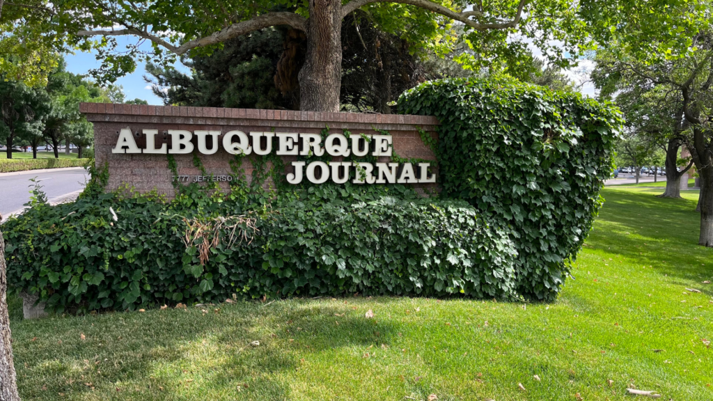 Sign reading "ALBUQUERQUE JOURNAL" covered in ivy, surrounded by grass and trees.