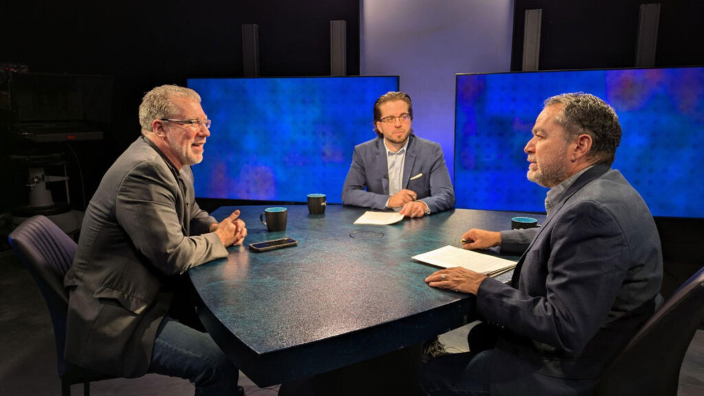 Three men in suits sit around a table in a TV studio setting, with cups and papers in front of them. Two large screens are visible in the background.