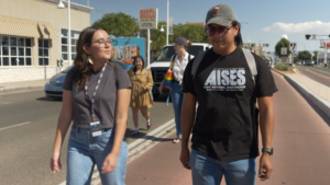 Two people walking on a sidewalk, both wearing casual clothing and glasses. The person on the right wears a cap. Other people are in the background, and shops line the street.