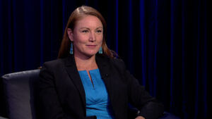 A person with long hair is seated in a chair, wearing a blue dress and a black blazer, against a dark curtain backdrop.