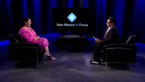 Two people are seated in a studio setting for an interview. The backdrop features a screen with the text "New Mexico in Focus.