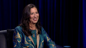 A woman with long brown hair, in a green and gold outfit, smiles while seated against a dark curtain backdrop.