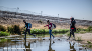 Three people with backpacks cross a shallow, rocky stream near a barbed-wire fence under a clear sky.