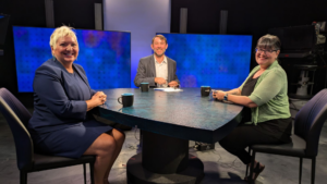 Three people seated at a round table in a studio setting with cameras and large screens in the background. They are smiling and have coffee mugs in front of them.