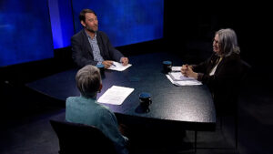 Three people are seated around a table in a dimly lit room with blue lighting. They have papers and mugs on the table, engaged in discussion.