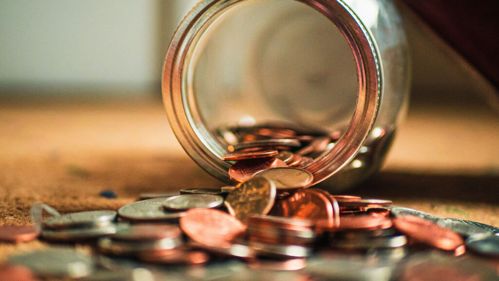 Coins spilling out of a tipped-over glass jar onto a wooden surface.