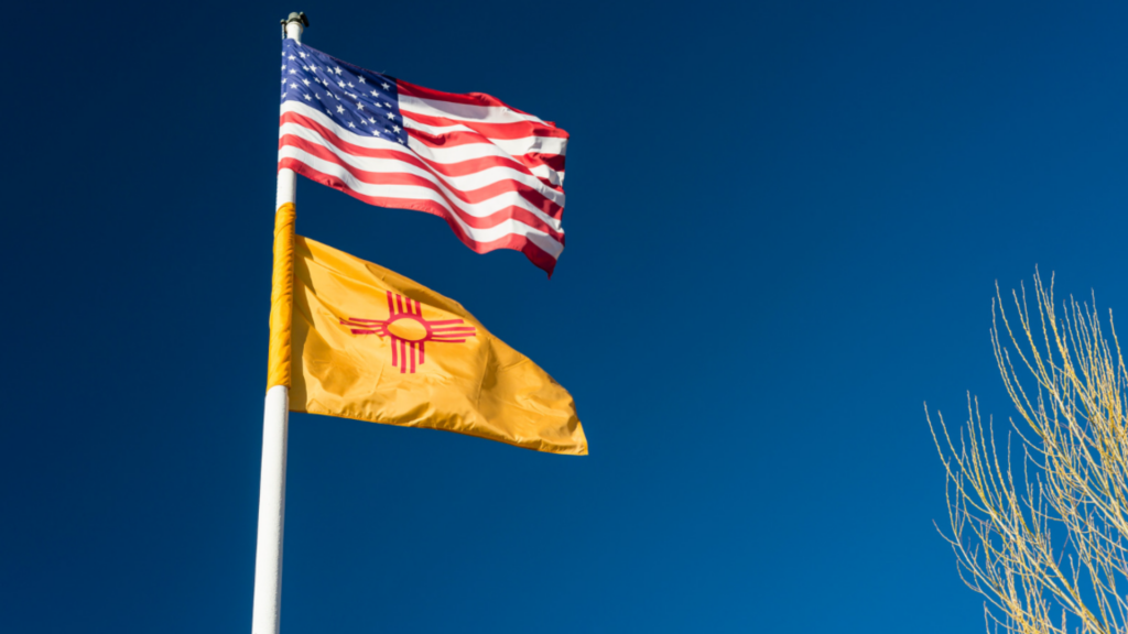 American and New Mexico flags flying on a pole against a clear blue sky.