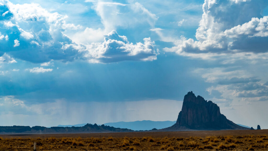 A rocky outcrop rises in a vast desert landscape under a partly cloudy sky with distant mountain ranges.