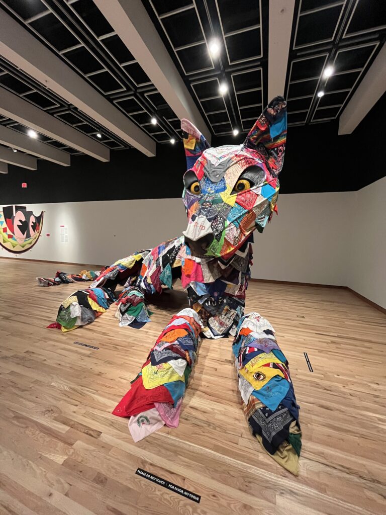 Large patchwork sculpture of a wolf, crafted from colorful fabric pieces, is whimsically displayed in a gallery with wooden floors. The atmosphere is fluid and dynamic, reminiscent of rippling water reflecting off the black ceiling above.