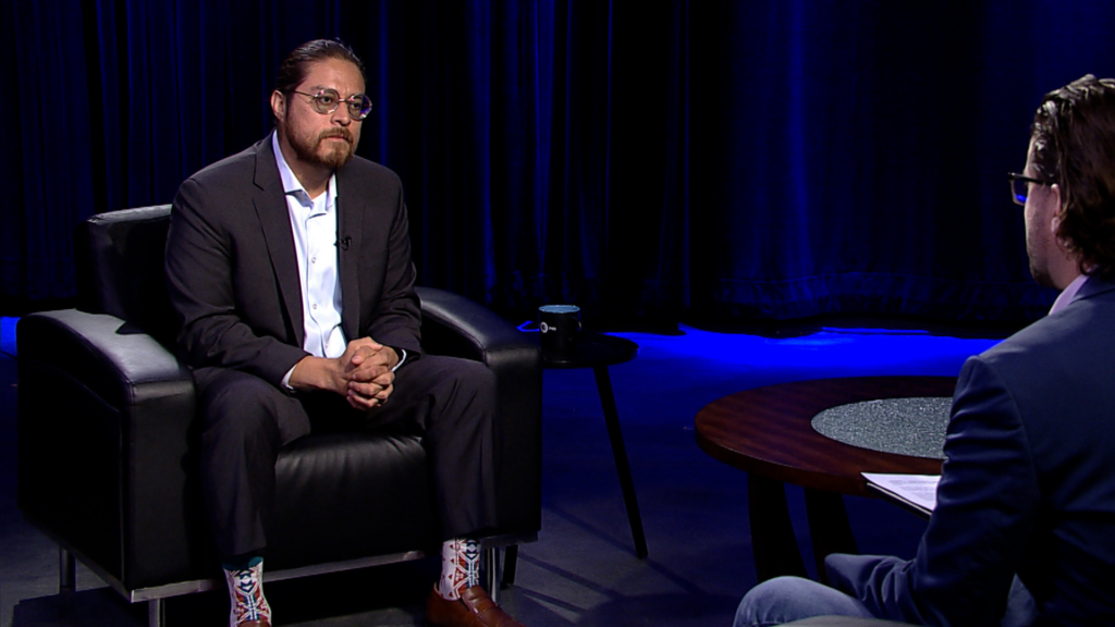 Two men seated in a studio setting with blue lighting, engaged in a conversation. One is on a black armchair, facing the other, who is partially visible from behind.