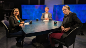 Three people sit at a round table in a studio, each with a cup in front of them, against a backdrop of large screens displaying blue patterns.