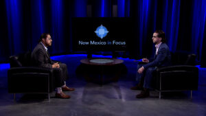 Two individuals seated in a studio setting with "New Mexico in Focus" displayed on a screen between them.