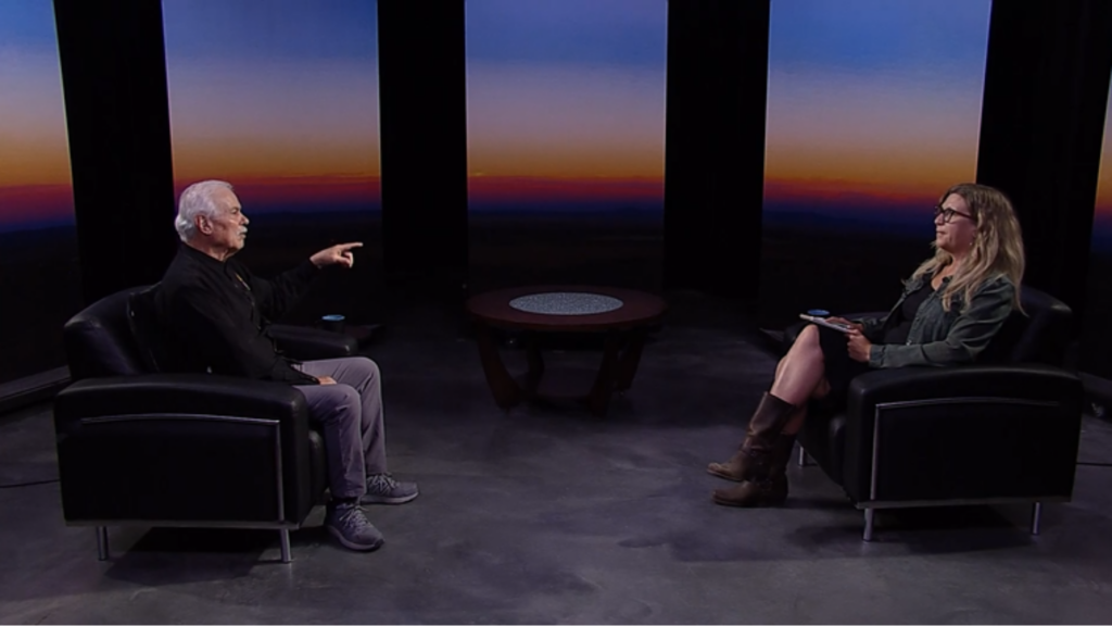 Two people sit in armchairs facing each other in a studio with a sunset backdrop. The person on the left gestures while the person on the right holds a notebook.