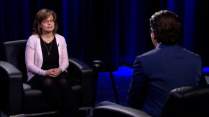 Two people seated facing each other in a studio with blue lighting. One wears a light pink cardigan, and the other is in a blue suit.