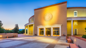 New Mexico State Capitol building exterior at dusk with illuminated entrance and decorative state seal.