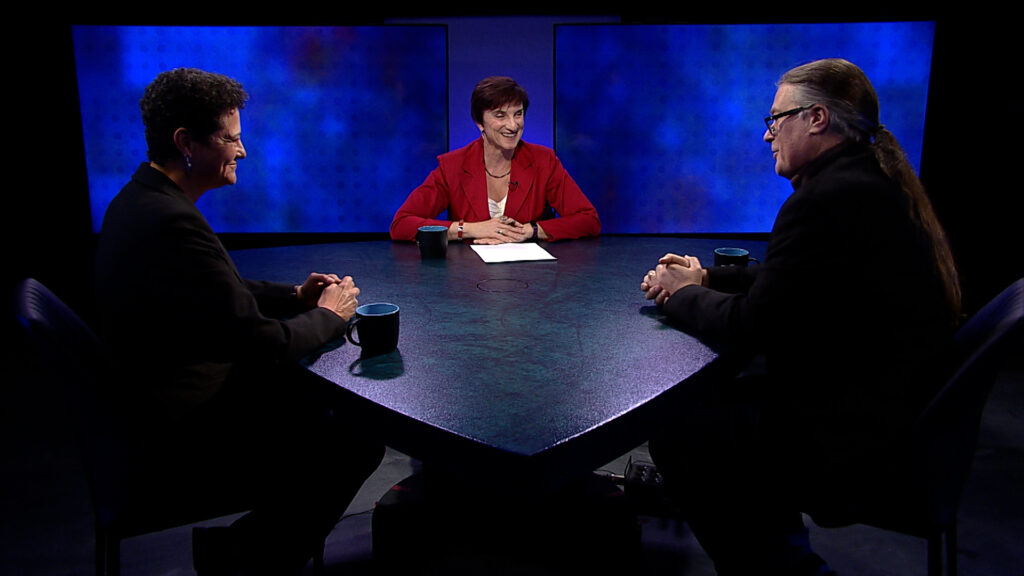 Three people seated at a triangular table in a discussion on a television set, with blue screens in the background.