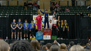 A group of students on a podium at a competition, with three top winners on the central platforms and others standing on the sides with medals.