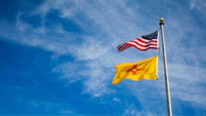 Two flags on a pole against a blue sky: the U.S. flag above a yellow flag with a red symbol, representing New Mexico.