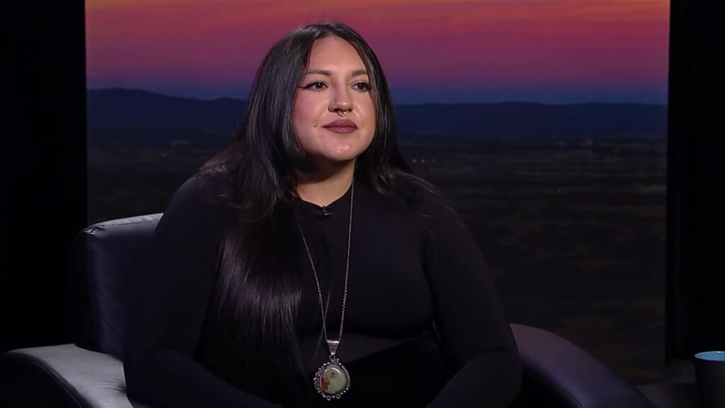 A person with long hair and a pendant necklace sits on a chair, gazing over the serene water, as the sunset paints the landscape in vibrant hues.