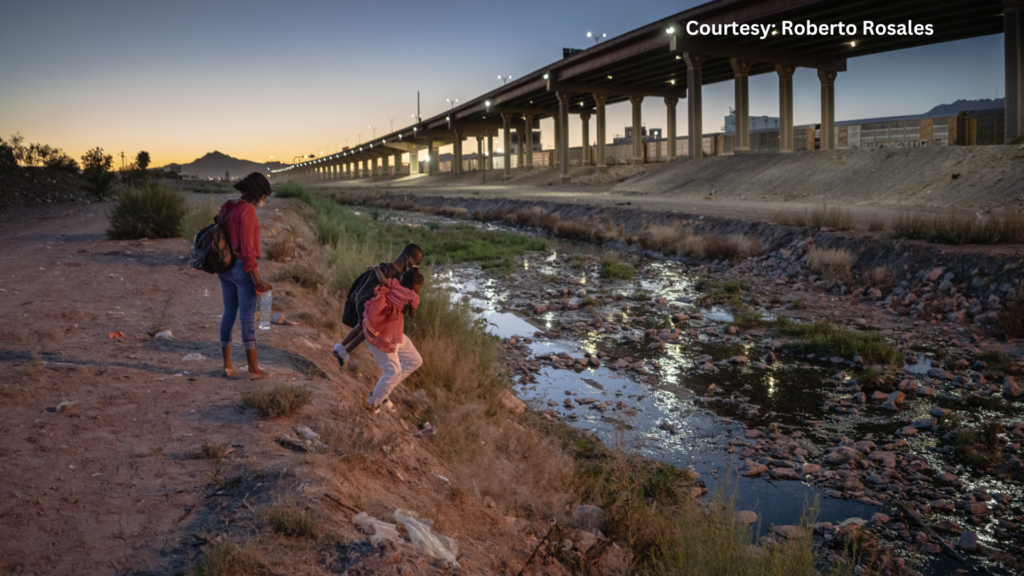 Two people stand at a riverbank at sunset, with one child stepping closer to the water. A bridge and a cityscape are in the background.