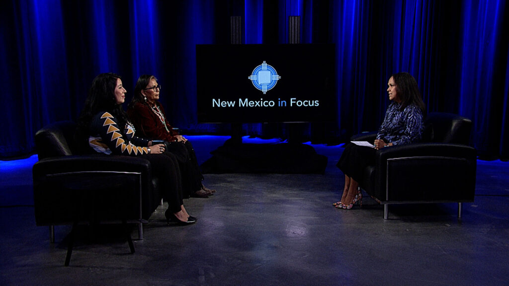 Three people are seated in a studio setting with a "New Mexico in Focus" screen. Two on one side, one on the other, under blue lighting.